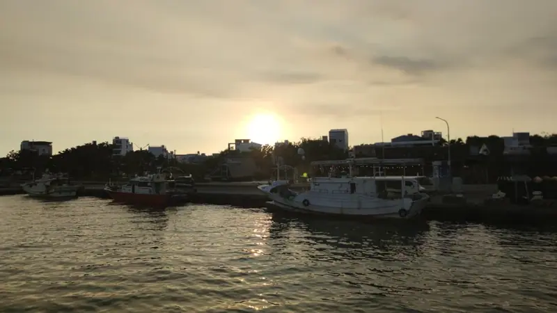 夕陽映照水面，從船上看岸邊停泊的漁船與建築