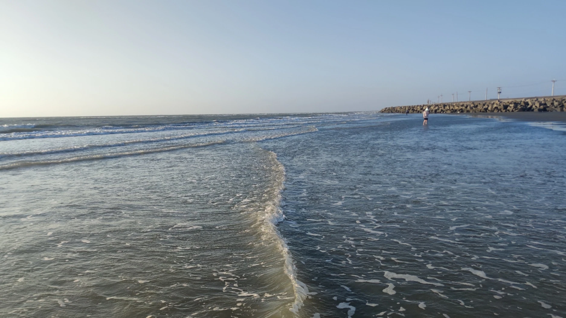 海浪沿著寬闊濕沙灘推進，遠處有人站在淺水邊