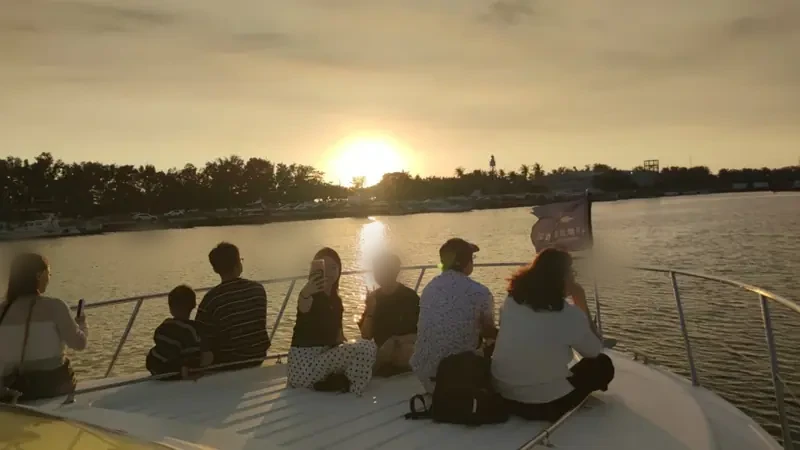 夕陽映照港面，幾位遊客坐在遊艇船首，其中有人拿手機自拍